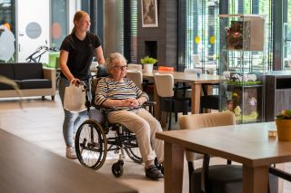 Zorgmedewerker duwt vrouw in een rolstoel door het restaurant in ZINN locatie De Es